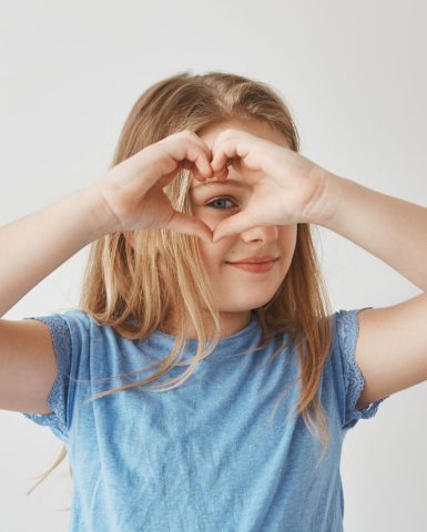 Close up of beautiful blonde girl making heart with hands, looking through it in camera, posing for photo with smile and happy expression